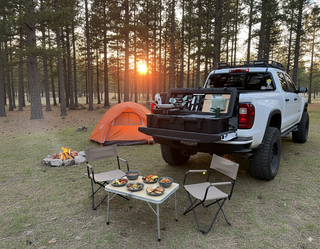 VOZ portable camp kitchen set up on a pickup truck tailgate at a forest campsite with tent and outdoor dining area